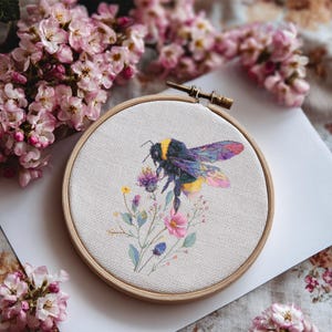 May include: A cross-stitch embroidery hoop with a bumblebee design. The bee, with yellow and black markings and iridescent wings, is depicted above colorful wildflowers. The embroidery is set in a wooden hoop, surrounded by pink blossoms.