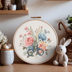 Pode incluir: Um bastidor de bordado em ponto cruz com um desenho floral e uma tesoura. As flores são em tons de rosa, azul e amarelo. A tesoura tem cabos azuis e está colocada sobre as flores. O bastidor é de madeira.