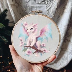 May include: A white dragon with pink and blue wings sits on a branch with pink flowers. The dragon is embroidered on a light gray fabric in a round wooden embroidery hoop.