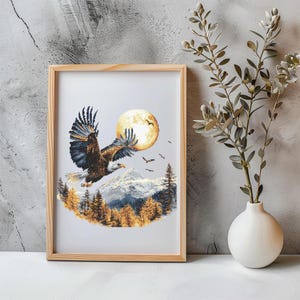 Puede incluir: Una bordado de punto de cruz enmarcado que representa un águila calva volando frente a una luna llena y una cordillera. El águila es marrón y negra con plumas blancas. La luna es amarilla y las montañas son blancas con árboles marrones.