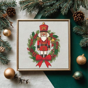 Può includere: Un ricamo incorniciato di un soldato schiaccianoci in uniforme rossa, circondato da una ghirlanda natalizia con bacche rosse e un grande fiocco rosso. L'opera è ambientata su uno sfondo festivo con rami di pino e ornamenti.