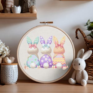 May include: A cross-stitch embroidery hoop featuring three Easter bunnies with pastel-colored egg bodies. Each bunny has a different patterned bow and ears. The embroidery is set in a wooden hoop, perfect for Easter decor.