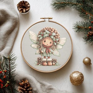 Könnte beinhalten: Ein Kreuzstich-Stickrahmen mit einem Feen-Design. Die Fee hat einen Kiefern- und Stechpalmen-Kopfschmuck, Flügel und einen roten Schal. Die Stickerei ist auf einem hellgrauen Stoff angebracht, umgeben von Tannennadeln, Tannenzapfen und goldenen Kugeln.
