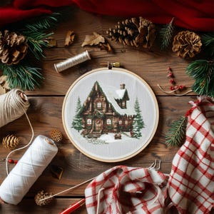 May include: A wooden embroidery hoop with a cross-stitch design of a snowy cabin with a wreath on the door. The hoop is surrounded by pine branches, pine cones, and red and white yarn.