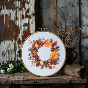 Può includere: Un cerchio da ricamo bianco con un design a corona autunnale. La corona è composta da pigne marroni, foglie arancioni e gialle e bacche rosse.