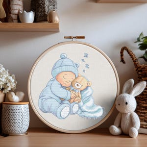 Può includere: Un ricamo a punto croce in un telaio di legno raffigura un bambino che dorme in pigiama e cappello blu, che abbraccia un orsacchiotto. L'orso è avvolto in una coperta blu. Lo sfondo è un tessuto beige chiaro. La parola "Zzz" è ricamata sopra la testa del bambino.