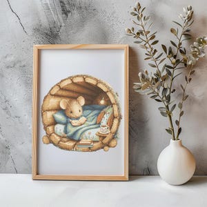 May include: A framed print featuring a cozy illustration of a mouse nestled in a log-cabin bed, reading a book. The mouse is under a blue blanket, with a small table holding a cup and books. The frame is light brown.