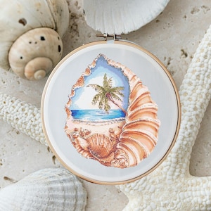 May include: A cross-stitch embroidery hoop with a seashell design. The seashell is open to reveal a beach scene with a palm tree and blue ocean.