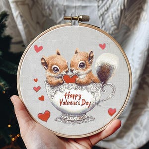 Könnte beinhalten: Zwei braune Eichhörnchen halten ein rotes Herz in einer weißen Teetasse mit einer Spitzenverzierung. Die Teetasse trägt den Text "Happy Valentine's Day". Der Hintergrund ist ein hellgrauer Stoff mit roten Herzen.