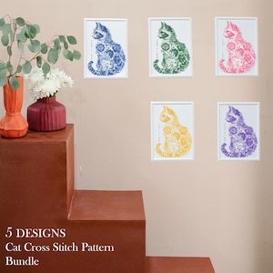 May include: Five cross stitch patterns featuring a cat design in different colors: blue, green, pink, yellow, and purple. Each pattern is framed and displayed on a wall.