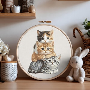 May include: A circular wooden embroidery hoop containing a cross-stitch pattern of three stacked kittens. The top kitten is calico, the middle is orange tabby, and the bottom is a gray tabby. The design is stitched on white fabric.