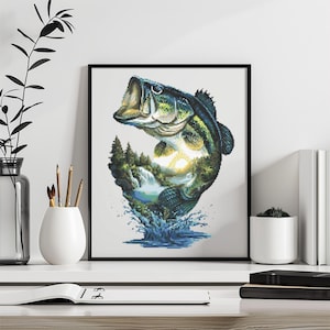 May include: A framed cross-stitch art piece featuring a large-mouth bass leaping from water. The fish's body contains a landscape of trees, a waterfall, and a sunset. The artwork is in a black frame and is displayed on a white surface.