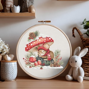 May include: A circular wooden embroidery hoop containing a cross-stitch design of a small brown mouse wearing a red knit hat and scarf, sleeping under a large red and white spotted mushroom, surrounded by pine branches, berries, and pinecones.