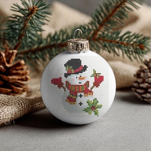 Puede incluir: Adorno navideño blanco con un diseño de punto de cruz de un muñeco de nieve. El muñeco de nieve lleva un sombrero negro, una bufanda roja y sostiene mitones rojos. El adorno tiene una tapa de metal dorado y se muestra con agujas de pino y una piña.