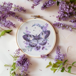 May include: A purple butterfly with a crescent moon and stars embroidered on a white fabric in a wooden embroidery hoop. The hoop is surrounded by purple lilac flowers.