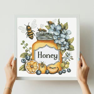 Peut inclure: Broderie au point de croix d'un pot de miel avec le mot "Honey" sur une étiquette blanche. Le pot est entouré d'abeilles, de myrtilles, de tranches d'orange et de fleurs bleues. L'œuvre est encadrée dans un cadre carré blanc.