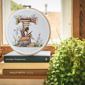 May include: A white embroidery hoop with a cross-stitch design of a girl reading a book on a stack of books. The design is surrounded by flowers and leaves. The hoop is sitting on a stack of books with the titles "Javier Marias", "Valkoinen Sydan", "Tanssikon Emile", "Philip Roth", and "Immisen Tahra".