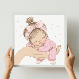 Può includere: Un disegno a punto croce di una bambina che dorme in un body rosa e una fascia abbinata con un fiocco. Il bambino è appoggiato su una mano grande, circondato da piccole forme di stelle. L'opera è incorniciata in una cornice quadrata bianca.