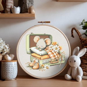 May include: A cross-stitch embroidery hoop featuring a sleeping mouse nestled in a suitcase. The suitcase is open, filled with blankets, flowers, and small boots. The color palette includes soft greens, oranges, and whites, creating a cozy, whimsical scene.
