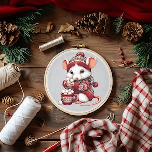 Puede incluir: Un bastidor de bordado con un ratón blanco con un jersey, gorro y bufanda rojos y verdes, sosteniendo una taza de malvaviscos. El bordado está sobre un fondo gris claro, rodeado de suministros de artesanía y decoración navideña.