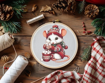 Op de afbeelding: Een borduurring met een schattige witte muis in een rood en groen outfit, die een mok marshmallows vasthoudt. De borduursel staat tegen een lichtgrijze achtergrond, omringd door knutselspullen en kerstversiering op hout.