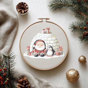 May include: A cross-stitch embroidery hoop featuring Santa Claus and a penguin in front of an igloo, surrounded by wrapped gifts. The design is set against a light gray fabric, framed by a wooden hoop. Festive holiday decor.