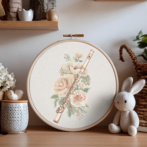 Könnte beinhalten: Kreuzstich-Stickerei in einem Holzrahmen mit einem Flötendesign und floralen Akzenten in zarten Rosa- und Cremetönen. Die Stickerei ist vor einem weißen Hintergrund angeordnet und wirkt zart und künstlerisch. Der Rahmen hat einen Durchmesser von etwa 20 cm.