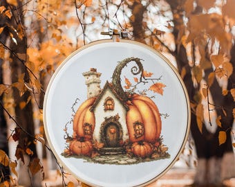 May include: A cross-stitch embroidery hoop with a whimsical autumn scene. A small house is built into a large orange pumpkin with a twisting vine growing from the top. Two smaller pumpkins are on the ground in front of the house.