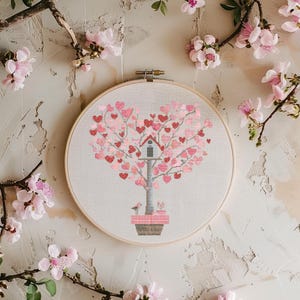May include: A cross-stitch embroidery hoop featuring a tree-shaped design made of pink and red hearts. The tree has a birdhouse and is set in a pot. The background is a neutral color, and the image is surrounded by pink flowers.