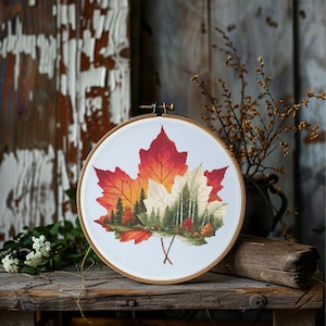 Op de afbeelding: Een borduurring met kruissteek met een herfstbladontwerp. Het blad toont tinten rood, oranje en geel, met een gedetailleerde bosscène in het midden. De borduursel zit in een houten ring.