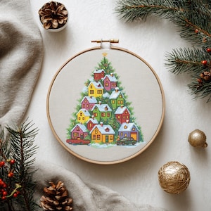 Puede incluir: Un bastidor de bordado de punto de cruz con un diseño de árbol de Navidad hecho de casas coloridas con tejados cubiertos de nieve. Las casas son rojas, amarillas, verdes y azules. Un coche rojo y un coche verde están aparcados en la base.