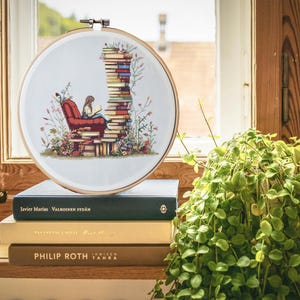 Peut inclure: Un cercle de broderie au point de croix représentant une personne lisant dans un fauteuil rouge, entourée de fleurs et d'une pile de livres. La broderie est fixée dans un cercle en bois. La scène se déroule devant une fenêtre.