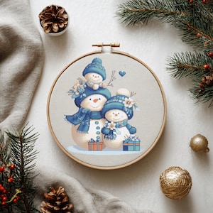 May include: A cross-stitch embroidery hoop featuring a family of snowmen. The snowmen wear blue hats, scarves, and hold gifts. The embroidery is set against a neutral fabric, with pine needles and ornaments in the background.