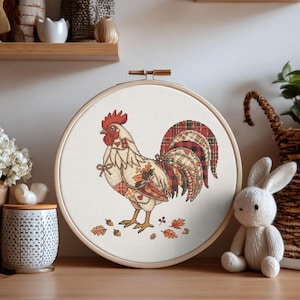 May include: A circular wooden embroidery hoop containing a cross-stitch design of a rooster. The rooster features a white body with red, orange, and brown plaid patterned feathers on its tail and wings, surrounded by small embroidered autumn leaves.
