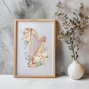 May include: A framed cross-stitch art piece featuring a harp design. The harp is beige and gold, adorned with floral accents in shades of cream and green. The artwork is displayed in a light wood frame, set against a neutral background.
