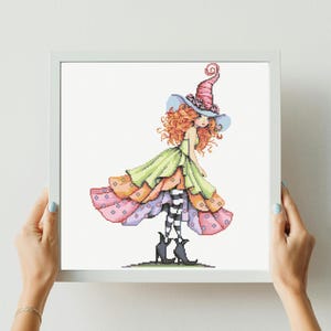 May include: A framed cross-stitch art piece featuring a whimsical witch. The witch has red curly hair, a pink and blue hat, and a layered dress in shades of green, orange, and pink. Her legs are striped black and white, and she wears black boots.
