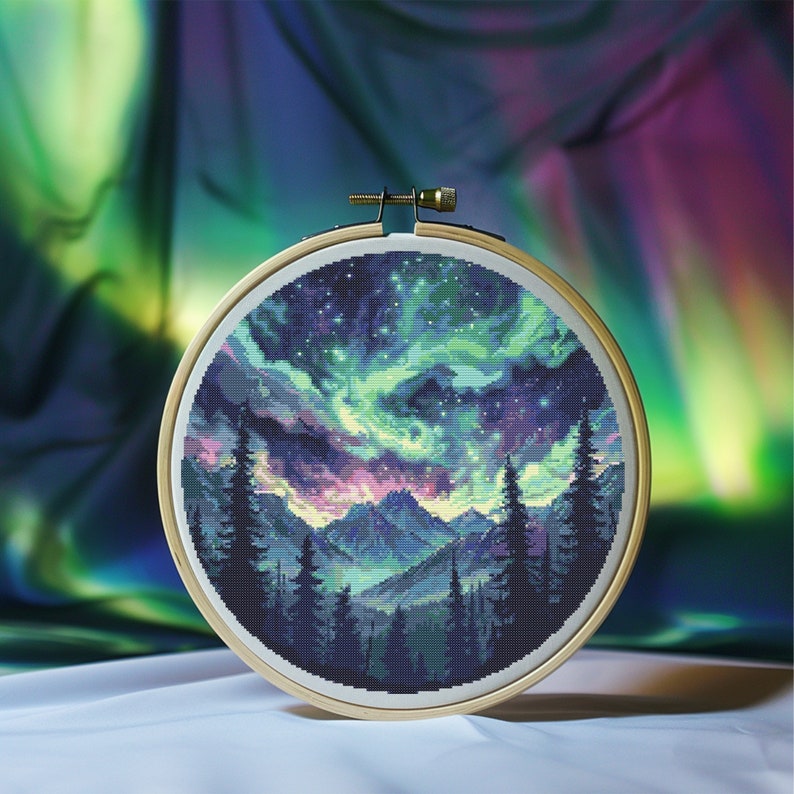 Puede incluir: Un aro de bordado de punto de cruz con un dise&ntilde;o de una cordillera de monta&ntilde;as bajo un cielo estrellado con una aurora boreal verde, morada y rosa.