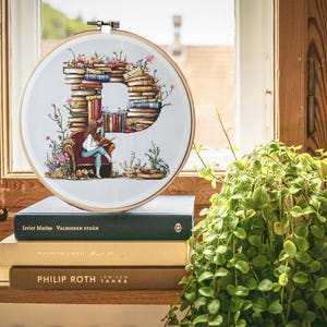 Może przedstawiać: Obręcz do haftu krzyżykowego z białym tłem i kolorowym wzorem litery P wykonanej z ułożonych książek. Mała figurka osoby siedzi na krześle i czyta książkę. Obręcz leży na stosie książek z tytułami "Javier Marias", "Valkoinen Sydan", "Tanssikon Emile", "PHILIP ROTH", "Immisen Tahra". W tle znajduje się zielona roślina.