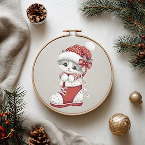 Pode incluir: Um bastidor de bordado em ponto cruz com um gatinho fofo usando um gorro e cachecol de Papai Noel, dentro de uma bota de Natal vermelha. O gatinho é cinza e branco, com um gorro e bota de bolinhas vermelhas e brancas. O bastidor é cercado por pinheiros e enfeites de Natal.