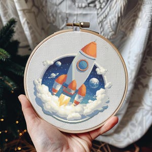 Op de afbeelding: Een borduurring met kruissteek met een raket die de ruimte in lanceert. De raket is wit en oranje, met blauwe en witte wolken en een sterrenachtergrond. De ring wordt in een hand gehouden.