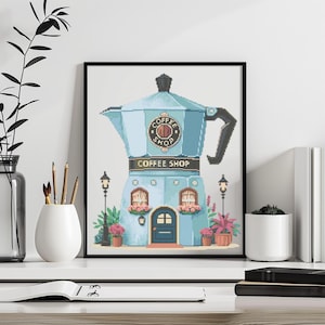 Puede incluir: Una obra de arte en punto de cruz enmarcada que representa una cafetería con forma de cafetera azul. La tienda tiene una puerta, ventanas y las palabras "Coffee Shop" están en exhibición. La obra de arte está rodeada por un marco negro.