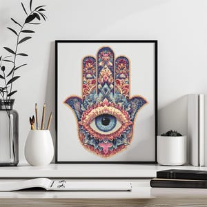 May include: Framed artwork featuring a detailed Hamsa hand design with an eye at the center. The artwork uses a pixelated style with a color palette of blue, pink, orange, and gold. The frame is black.
