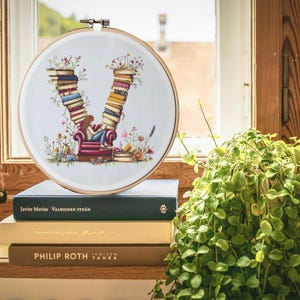 Könnte beinhalten: Ein weißer Stickrahmen mit einem Kreuzstich-Design des Buchstabens V, der aus gestapelten Büchern besteht. Eine kleine Figur sitzt auf einem roten Sessel und liest ein Buch. Der Rahmen liegt auf einem Stapel Bücher mit den Titeln "Javier Marias", "Valkoinen Sydan", "Tayson Emily", "Philip Roth" und "Immisen Tahra".
