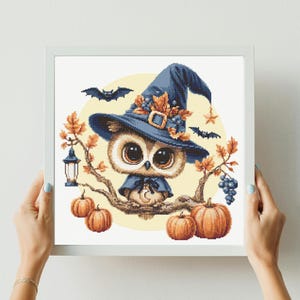 Può includere: Uno schema a punto croce incorniciato raffigurante un gufo beige con un cappello da strega blu decorato con foglie arancioni. Il gufo è appollaiato su un ramo con zucche arancioni, una lanterna, uva viola e due pipistrelli neri su sfondo giallo.