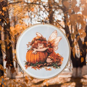 Puede incluir: Un aro de bordado de punto de cruz con un fondo blanco que presenta una hada con alas naranjas y un vestido marrón durmiendo sobre una gran calabaza naranja. El hada tiene el pelo marrón con una corona de bayas rojas y hojas. La calabaza está rodeada de hojas marrones y bellotas.