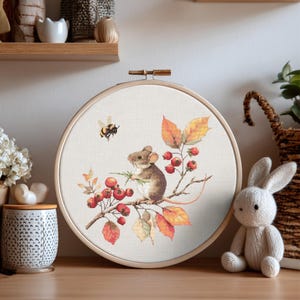 Könnte beinhalten: Ein Kreuzstich-Stickrahmen mit einer Maus auf einem Ast mit roten Beeren und Herbstblättern. Eine Biene fliegt in der Nähe. Das Design ist in einem Holzrahmen gefasst.