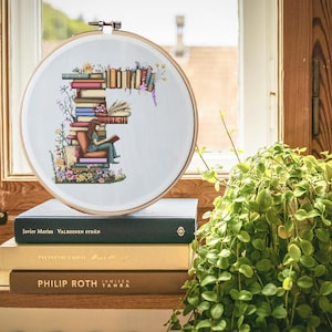 Può includere: Un cerchio da ricamo bianco con un disegno a punto croce della lettera F fatto di libri impilati. Una piccola figura è seduta sui libri a leggere un libro. Il cerchio è appoggiato su una pila di tre libri.