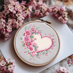Peut inclure: Un cercle de broderie au point de croix avec un motif de cœur rempli de fleurs roses et de feuilles vertes. Le cœur est souligné en rose et entouré d'un cercle en bois. Des fleurs roses sont disposées autour du cercle.