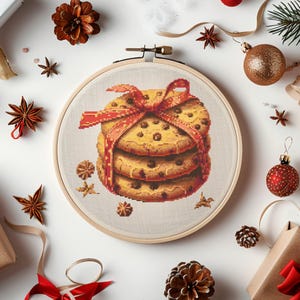 Könnte beinhalten: Nahaufnahme eines Kreuzstich-Stickrahmens mit einem Stapel von drei Schokoladen-Chip-Keksen, die mit einem roten Band zusammengebunden sind. Die Kekse befinden sich auf einem beigefarbenen Stoffhintergrund.