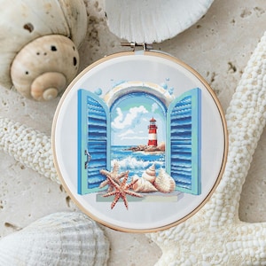 May include: A white embroidery hoop featuring a blue and white window scene. The window is open, revealing a view of a red and white lighthouse on a rocky shore with blue water and white waves. Seashells and a starfish are placed on the windowsill.
